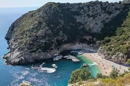 Vista aérea de la Bahía de Grama en Vlora, Albania, con lanchas rápidas fondeadas en aguas turquesas y visitantes descansando en la playa de guijarros blancos.